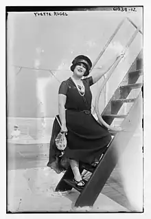 A young white woman standing on stairs on the deck of a ship, smiling, wearing a dark hat and dress, and strings of pearls.