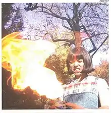 A photograph of an angry woman with short black hair wearing a flannel shirt and overalls. She is holding a compressed air can which is spewing flames that she is glaring at. She is stood in front of a row of trees in daytime. There is a narrow plain white border around the image where the name of the album is handwritten in the top-right corner.