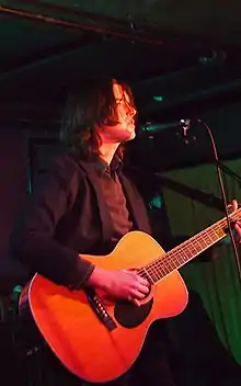 Young man with shaggy hair, on stage singing into a standing mic while playing the guitar