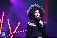 Zainab Johnson has brown skin, and an afro. She is wearing a black top and appears to be on stage.