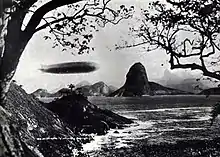 A black-and-white photograph taken between two trees. The Graf Zeppelin is flying at low level from left to right above a rocky coastline. In the background is the distinctive point known as Sugar Loaf, which marks the entrance to Rio de Janeiro harbour.