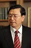 a serious looking man with a wavy haircut, wearing glasses, a white shirt, a suit and a red tie with white stripes and looking leftwards (away from the camera)