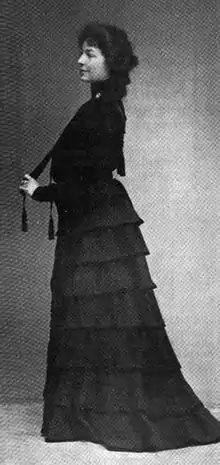 A young white woman, standing in profile, wearing a long dark dress with a tiered skirt