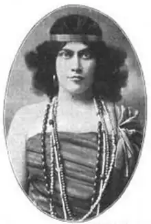 A light-skinned woman with dark hair and eyes, wearing a costume involving a headband, a strapless bodice, and beads. She is square to the camera, and the photo is in an oval frame.