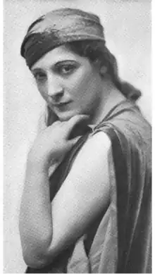 A white woman with dark eyes, chin in hand, arm bare to shoulder, wearing a fabric headwrap and a loose-fitting garment
