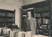 Young Hendrik Coenraad Blöte standing in an office surrounded by books. Hendrik is wearing a suit, tie, waistcoat and jacket.