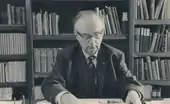 Older Hendrik Coenraad Blöte working at his desk. Hendrik is wearing glasses. a suit, tie and jacket. Many reference books can be seen on shelves in the background.