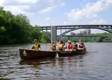 Seven young people in canoe, shoreline is green, women paddling, all wearing life vests, bridge span and university visible behind them