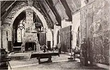 A monochrome photograph of a grand living room with a 36-foot-high arched ceiling, a high fireplace, and tall stained glass windows.