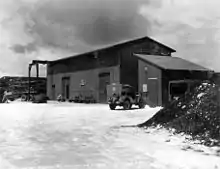 Two buildings with sloped roofs. Outside is parked a jeep and a truck.