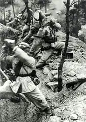 Una fila de soldados está de pie frente a una trinchera y sostiene piedras sobre sus hombros. Las rocas vuelan hacia la derecha mientras el humo llena el fondo.