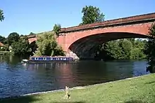 Un ancho río arbolado a la izquierda tiene un banco verde en el primer plano a la derecha y está atravesado por un puente bajo de dos arcos de ladrillo. Un árbol en medio del río oscurece parte del puente.