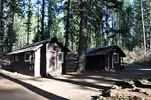 Two small cabins in the woods face a dirt road.