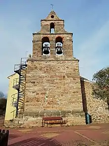 Campanar de la iglesia del pueblo vista desde una perspectiva frontal. En la parte más alta tiene dos campanas. Es de piedra y contrasta con la parte renovada que se ve.
