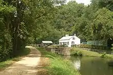  Fotografía de un camino de sirga de tierra junto a un canal con una esclusa con una casa al fondo