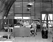 An office with desksm chairs and boxes inside a semi-cylindrical corrugated iron building.