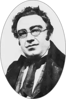 Round-faced white man, clean shaven with unruly dark hair and round spectacles