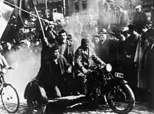 Trois jeunes hommes sur un side-car, brandissant un drapeau ; un attroupement en arrière-plan.