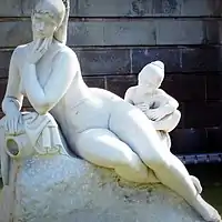 Escultura de piedra blanca que representa a una mujer sentada, recostada ligeramente sobre su lado derecho y una niña sentada. Ambas desnudastras ella