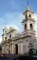 Frontis, Campanarios, Atrio y Galería lateral de la Catedral Basílica de Salta.Situada frente a la Plaza 9 de Julio, en el corazón del centro histórico de la Ciudad, esta Catedral integra un conjunto monumental con el Palacio Episcopal. Circunscribiéndonos a su parte más importante, en el año 1.873 con el fin de reanudar trabajos de construcción largamente paralizados, se contrató la construcción de la parte remanente y aún inconclusa, con la Empresa de los arquitectos Nicolás y Agustín Cánepa, excluyendo: frente, atrio, torres y galería al costado izquierdo desde la entrada a la sacristía. A partir de allí, pseudo investigaciones históricas, en algunos casos empeñados en deformar la verdad histórica sobre la autenticidad en la autoría del proyecto del frente, atrio y campanario, pretendieron y aun pretenden en esta ambigüedad, adjudicar su autoría a: ¿Bertrés?, ¿Soldati ?, ¿Georgi?, ¿Riguetti?, generando al día de la fecha, conclusiones erróneas fácilmente evitables. Un documento redescubierto por la Academia Nacional de Bellas Artes, merced a la búsqueda de documentación efectuada por la investigadora de campo, Sra. Iris Gori, bajo la supervisión del académico de número Prof. Héctor H. Schenone, restablece merced a este hallazgo de modo fehaciente, que el verdadero autor del diseño es el Sr. Maqui (apellido escrito alternativamente como Maqui o Macchi por el propio arquitecto Macchi, a fin de facilitar su pronunciación). A fs.271,el libro Patrimonio Artístico Nacional, Academia Nacional de Bellas Artes, Bs. As. año 1988, dejó consignado: «...Hemos podido levantar una interesante noticia en el Diario salteño «La Reforma» del 28 de junio de 1879 (pág. 2, 3era. columna): «...Por 29000 $ se ha contratado la obra del frontis de la Catedral, con el empresario Riguetti según el plano del Señor Maqui...».