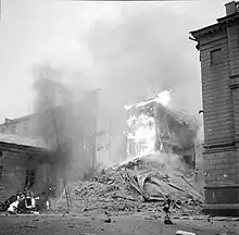 An apartment building is on fire and has partly collapsed in Central Helsinki after Soviet aerial bombing on 30 November 1939. A woman in a coat and a hat is passing on the right next to the rubble and a car is on fire on the left.