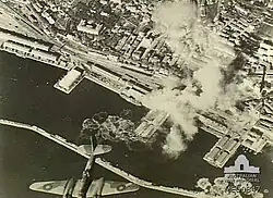 Vista aérea del puerto y columnas de humo causadas por explosiones con un bombardero al frente.