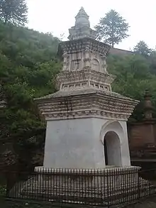 A two storied white pagoda with a fence surrounding it. In the background is a green hill.