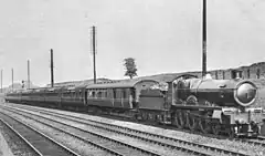 Una imagen en blanco y negro de cuatro líneas ferroviarias en un corte poco profundo, una gran máquina de vapor conduce un tren de autocares desde el centro de la izquierda hasta la derecha en primer plano