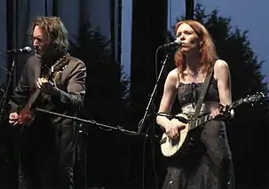 A middle-aged man wearing a dark suit playing guitar while Welch in a black striped dress plays a banjo and sings on stage.