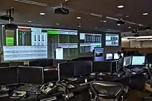 Una oficina oscura llena de computadoras; pantallas de datos financieros llenan una pared