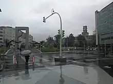 La imagen muestra La Plaza de América tras la remodelación con la Puerta del Atlántico enfrente mientras llueve y un transeúnte espera a cruzar la calle.