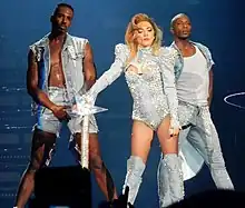 A blond woman in a nude colored leotard is flanked by two males in nude body suit and blue jacket. The woman's chest and the hand and head of the two males .