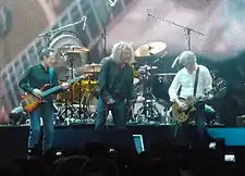 Led Zeppelin onstage: John Paul Jones playing bass guitar, Robert Plant holding a microphone, and Jimmy Page playing guitar. The trio stand in front of Jason Bonham's drum kit.