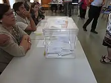 Una mesa electoral con una urna transparente, que contiene varios sobres.