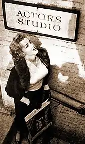 Monroe, que lleva falda, blusa y chaqueta, de pie debajo de un cartel del Actors Studio mirando hacia él.