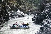 Two rafts, one with three people and one with one person, negotiate fast-moving water in a narrow, rocky river canyon.