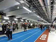 Pintura que simula pistas de atletismo dentro de un aeropuerto. En una pista, se pueden ver pictogramas que indican dónde ir para coger el metro o llegar al aparcamiento