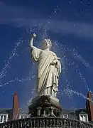 Statue blanche, sur fond de ciel bleu, encadrée par des jets d'eau