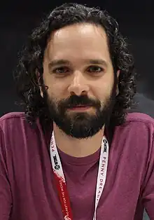 36 year-old man with curly black hair and a beard smirking at the camera.