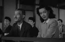 A theatre at which a Noh play is being performed: Prof. Somiya is wearing a business suit and tie and Noriko is wearing a simple, Western-style dress with a collar; Shukichi is looking straight ahead towards the left frame of the picture, smiling, and Noriko, not smiling, is looking toward the right frame of the picture towards an unseen person.