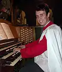 Fotografía de color de un organista sonriendo blanco y rojo tocando el órgano de St James'. La partitura, teclado, y algunas partes del órgano son visibles
