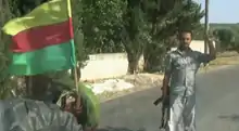 Una bandera con barras horizontales de color amarillo rojo y verde unidas a un vehículo militar (izquierda) al lado de un luchador kurdo YPG con un AK-47. (derecho)