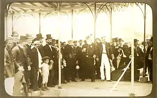 The white-bearded emperor stands among a group wearing formal and military attire under a pavilion.