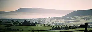 a panoramic view of green fields with a hill sloping up to the right. In the background is a lighter hill, rising gently from the right and dropping sharply on the left.