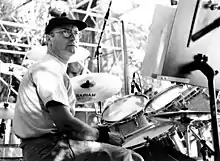 A middle-aged man, wearing a hat, glasses, light top, jeans and a wristbands, playing on a drumkit.