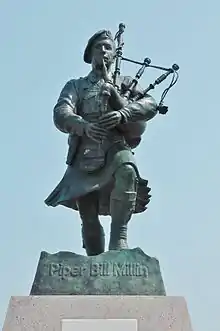 Fotografía de una estatua de bronce de tamaño natural con pátina verdigris que representa a Millin marchando con su falda escocesa y tocando la gaita