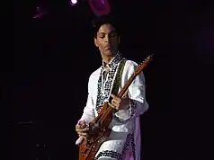 Prince tocando la guitarra con una camisa blanca con pedrería.