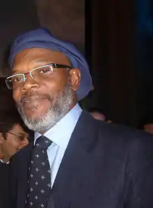 A middle-aged man, wearing a blue hat, glasses and a suit.