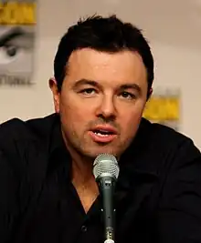 A Caucasian male with black hair and stubble, along with a black shirt. His hand is placed under his chin and a microphone is in front of him. A vague simple is marked behind him.