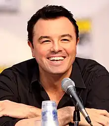 Un hombre de pelo corto negro y camisa negra, de piel bronceada, ríe por un micrófono mientras se inclina hacia adelante.